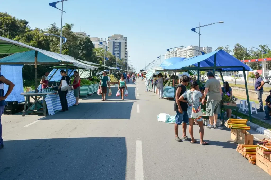 Street market on a sunny day with blue canopies and vendors selling goods. People walk along the road, flanked by trees and tall buildings.