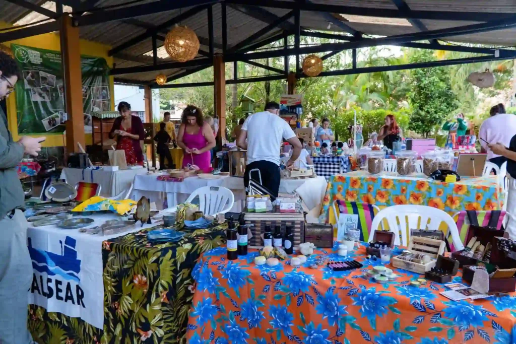 Outdoor market with vibrant, colorful tables covered in crafts, oils, and homemade goods. People browse under a rustic pavilion on a sunny day.