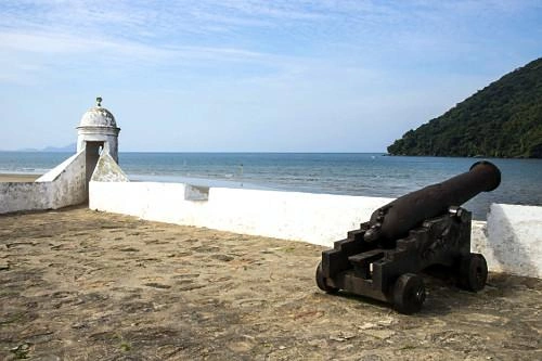 A weathered stone fortress terrace by the sea, featuring a white sentry box with a domed roof and an antique black cannon mounted on a wooden carriage, against a backdrop of ocean and distant mountains.