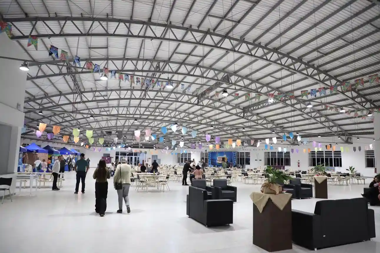 A wide, bright view inside a large, industrial-style event hall or pavilion. The space features a high ceiling with a visible metal truss framework and white insulation panels, adorned with strings of colorful triangular pennants. The floor is a glossy white. Scattered throughout the room are numerous white square tables with chairs, set up for dining or gatherings. In the foreground, there are black, modern armchairs and dark brown planters with greenery, draped with light fabric. Several attendees are walking and mingling across the floor.