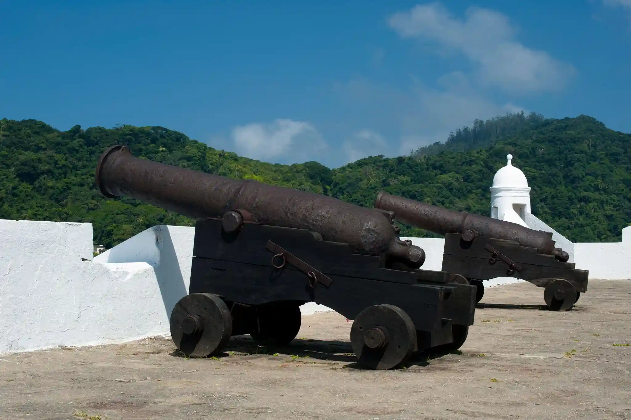 A wide view of a fort's stone terrace lined with multiple dark cannons pointing toward the water, bordered by a thick white parapet wall, with a sandy beach, calm blue bay, green mountains, and a tiled-roof building visible nearby.
