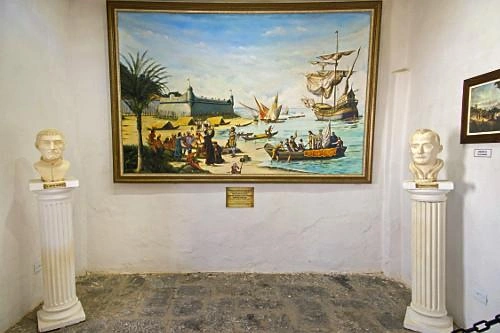 An indoor museum display inside a white-walled room. Two classical white marble busts stand on fluted columns flanking a large, framed historical painting. The painting depicts a coastal scene with a tall sailing ship, a smaller rowboat, and people on a sandy beach near a fortress with a palm tree. A small informational plaque is mounted on the wall below the artwork.