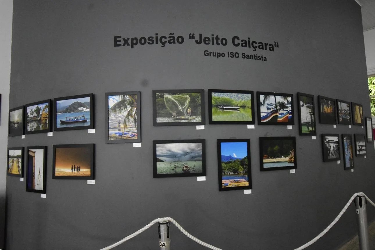 An indoor photography exhibition displayed on a matte dark gray wall. The text at the top, written in black, reads "Exposição 'Jeito Caiçara'" with a smaller subtitle "Grupo ISO Santista". Below the text, a selection of framed color photographs is arranged in two horizontal rows. The photos depict various coastal and nature scenes, including ocean views, palm trees, boats, and people on a beach. Small, white rectangular labels are placed beneath each frame. In the foreground, a physical barrier made of white rope strung between metal stanchions separates the viewer from the artwork.