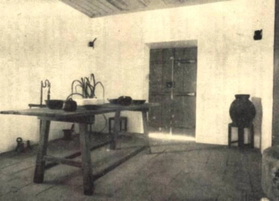 A vintage, sepia-toned photograph of a rustic room with bare, textured walls and a plank floor. In the center is a sturdy wooden table holding various items, including an old iron stand, a kettle, and bowls. Against the back wall is a heavy, dark wooden door with iron hinges, and to the right, a large dark jar rests on a small wooden stool.