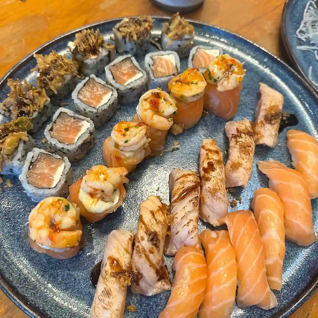 A blue plate with assorted sushi rolls, shrimp-topped pieces, and seared salmon nigiri, arranged neatly on a wooden table.