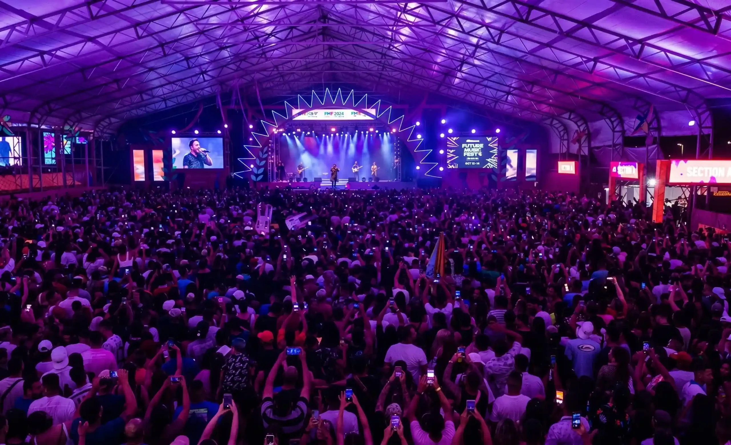 A large indoor music festival with thousands of people facing a brightly lit stage under purple lighting, many attendees holding up smartphones to record the live performance.