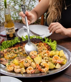 A person serves shrimp and potatoes from a platter with grilled fish. A Corona beer bottle is on the table beside a white plate.