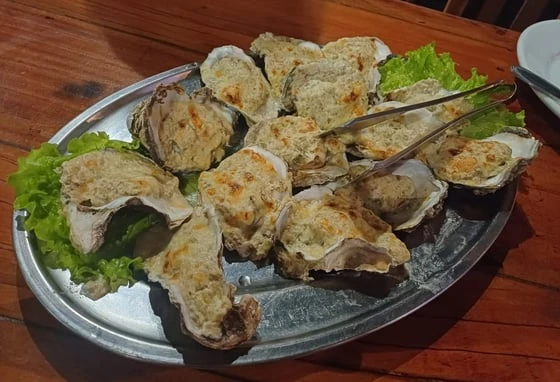Grilled oysters on a metal platter with lettuce garnish, served on a wooden table, accompanied by tongs for easy serving.