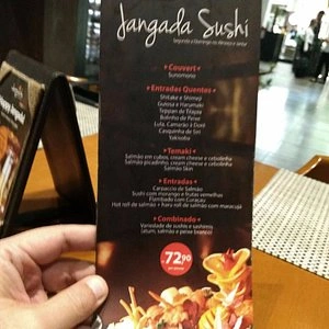 Hand holding a Jangada Sushi menu featuring various sushi options and a highlighted price of 72.90, set on a wooden table.
