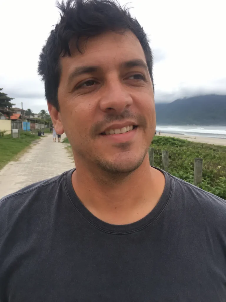 A man with short dark hair and a stubble beard stands on a paved walkway near the beach, with cloudy skies and mountains in the background.