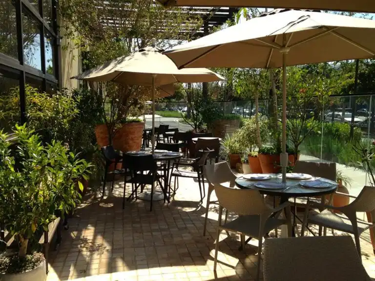 Outdoor cafe with tables, chairs, and umbrellas surrounded by lush greenery on a sunny day, creating a relaxing atmosphere.