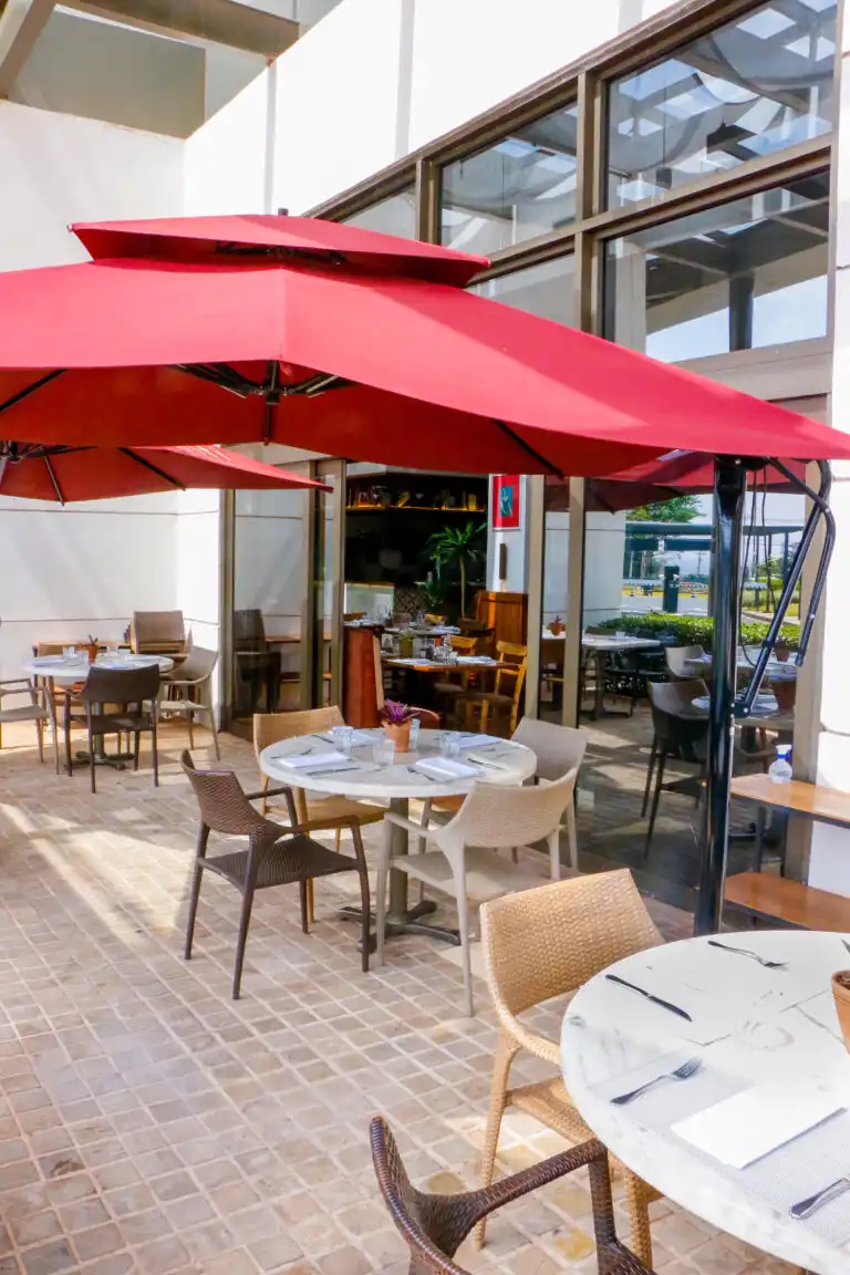 Outdoor restaurant seating with red umbrellas, round tables, and wicker chairs on a sunny patio, ready for dining guests.