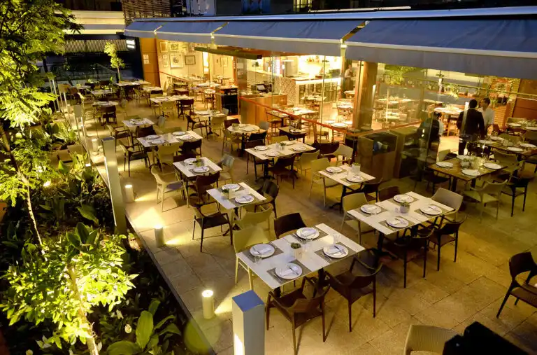 Outdoor restaurant with neatly set tables, glowing lights, and lush greenery, creating a warm, inviting dining atmosphere at night.