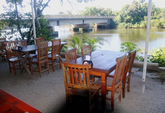 Outdoor riverside cafe with wooden tables and chairs, overlooking a bridge and lush greenery under a clear sky.