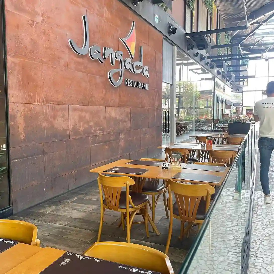 Outdoor seating at Jangada Restaurante with wooden tables and chairs, a person walking by, and a modern glass railing.