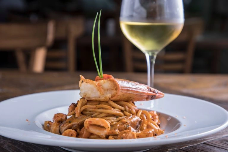 Seafood pasta topped with shrimp, garnished with chives, paired with a glass of white wine on a rustic wooden table.