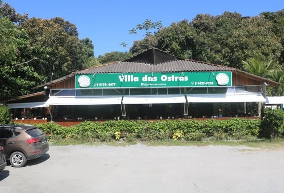 Villa das Ostras restaurant with a green sign, surrounded by lush trees, and parked cars in front on a sunny day.