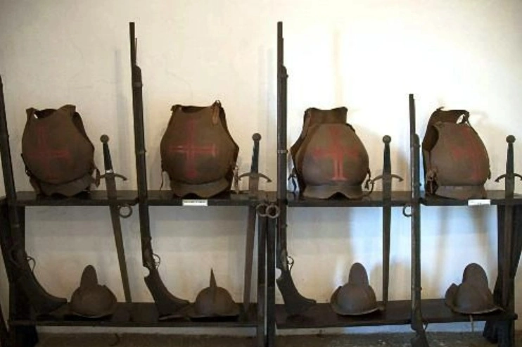 A close-up view of a two-tiered weapons rack displaying four brown breastplates marked with a red cross on the upper shelf, alongside muskets and swords, and several helmets arranged on the lower shelf.
