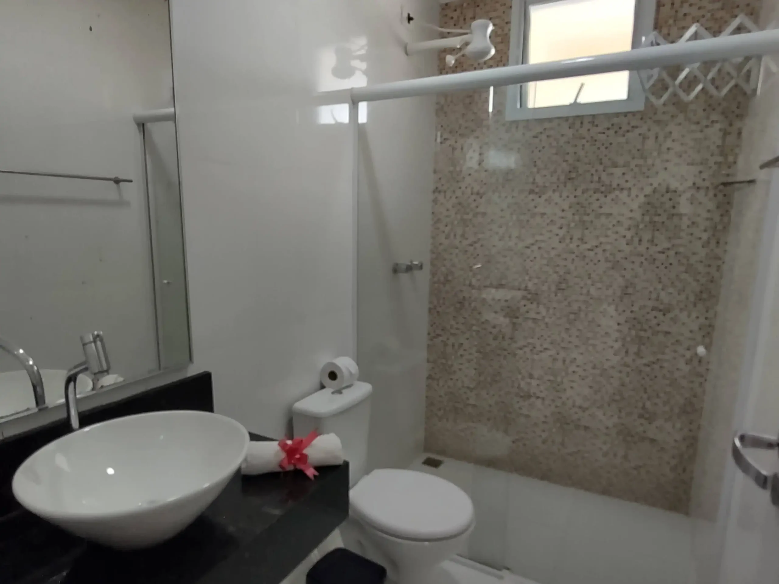 Clean, modern hotel bathroom featuring a white vessel sink on a black countertop, a large mirror, a wall-mounted hair dryer, and a walk-in shower with beige mosaic wall tiles and a high window.