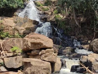 A natural waterfall flowing down a steep, rugged hillside composed of large, stacked, light-brown boulders and rock ledges, surrounded by scattered green vegetation and exposed tree roots.