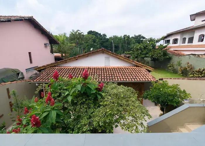 View of a pink residential building and a separate structure with a red clay tile roof, surrounded by lush green trees and tropical red flowering plants in a sunny backyard.