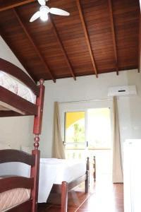 A cozy bedroom featuring a striking sloped ceiling made of dark wooden planks and a white ceiling fan, with a wooden bunk bed and a single bed dressed in crisp white bedding, illuminated by bright natural light.