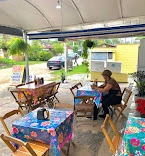 Open-air covered patio with wooden chairs, colorful floral tablecloths, a seated patron, and a yellow food stand in the background.
