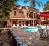 A sunny tropical swimming pool surrounded by white lounge chairs at a rustic, family-friendly pousada hotel with palm trees and a terracotta patio.