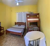 Sunlit, budget-friendly guest room with vibrant yellow walls, featuring a wooden double bed, a wooden bunk bed, a ceiling fan, and a small round dining table with two chairs on tiled flooring.