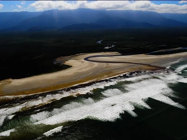 Wide aerial landscape of the sandy Itaguaré river estuary meeting the deep blue ocean, with white waves crashing against the shore and a majestic mountain range under a cloudy sky.