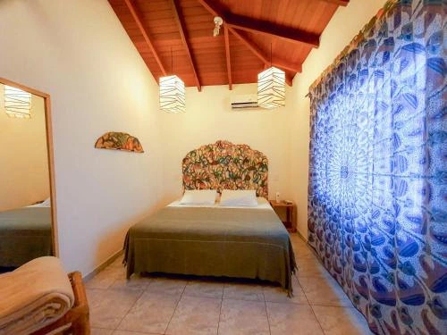 Comfortable tropical bedroom featuring a large bed with a dark green cover, floral patterned headboard, tall wood ceiling, and blue-and-white circular patterned curtains.