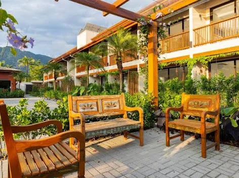 Inviting outdoor garden lounge area featuring sturdy wooden benches and chairs, surrounded by lush green tropical plants, purple flowering bushes, and a nearby resort building.