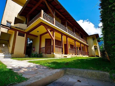 Exterior of a traditional yellow-painted hotel or lodge featuring prominent wooden structural beams, wrap-around balconies, and a landscaped front lawn under a bright sky.