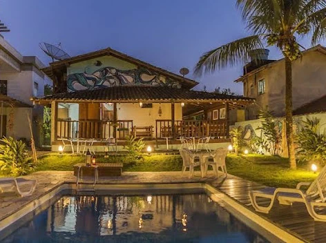 Evening scene of a residential or boutique hotel backyard featuring a small rectangular pool, white patio chairs, outdoor string lighting, and a house with a painted mural on the upper wall.