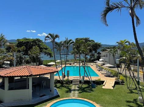 Large resort swimming pool surrounded by a green lawn, tall palm trees, and white umbrellas, offering a picturesque view of the nearby ocean and distant mountains.