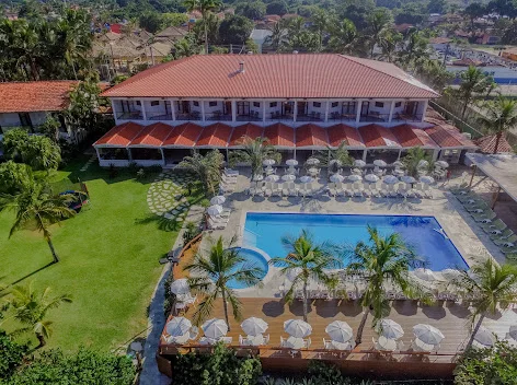 Bird's-eye aerial view of a resort hotel featuring a large outdoor swimming pool, a wide sun deck with numerous white umbrellas, and a large red-tiled main building.