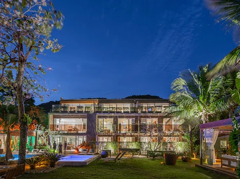 Luxury modern multi-story resort exterior illuminated at night, featuring glass-walled rooms, outdoor lounge areas, a small pool, and palm trees against a starry dark blue sky.
