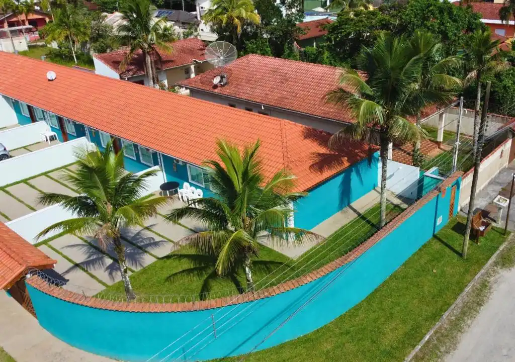 An elevated drone shot of a residential or vacation property featuring a vibrant curved turquoise boundary wall topped with barbed wire, a long building with red terracotta roof tiles, and several tall palm trees casting shadows on the grass and paved courtyard.