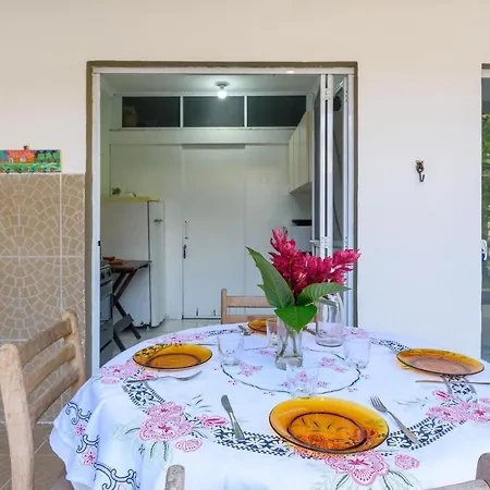 A round outdoor dining table set with yellow ceramic plates, silverware, and a vibrant red floral centerpiece, sitting on a white patterned tablecloth with the kitchen visible in the background.