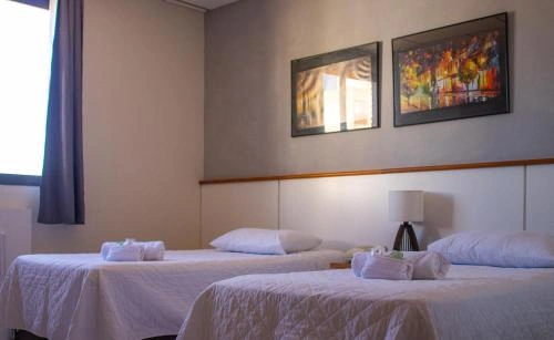 A neat, modern hotel room featuring two twin beds with bright white duvets and fresh white towels, a white headboard with a wooden trim, and two framed artworks hanging on the beige wall above a small bedside lamp.