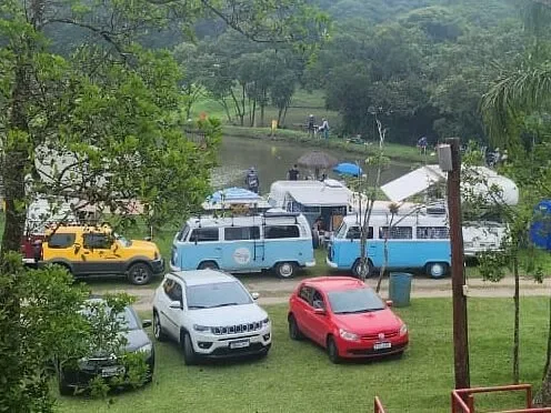 An outdoor gathering scene showing several vehicles parked on a grassy hill overlooking a calm lake, featuring a yellow SUV, a white SUV, a red sedan, and two classic blue and white vans, with groups of people visible near the water's edge.