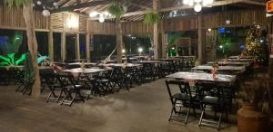 A spacious, open-air event venue at night featuring a high ceiling with exposed wooden beams, natural tree trunk pillars, and strings of globe lights. Long wooden tables with white tablecloths and black folding chairs are arranged in rows, illuminated by ambient warm lighting.