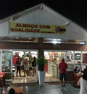Nighttime restaurant storefront with glass doors, red brick walls, dining patrons, and a yellow banner reading "Almoço com Qualidade".