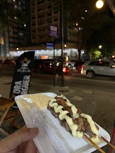 Hand holding a white tray with a meat skewer drizzled with white cream sauce, in front of a nighttime city street and a black burger stand flag.