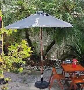 Large grey patio umbrella on a black stand, positioned over a wooden table with a red cup, surrounded by stone landscaping and green plants.