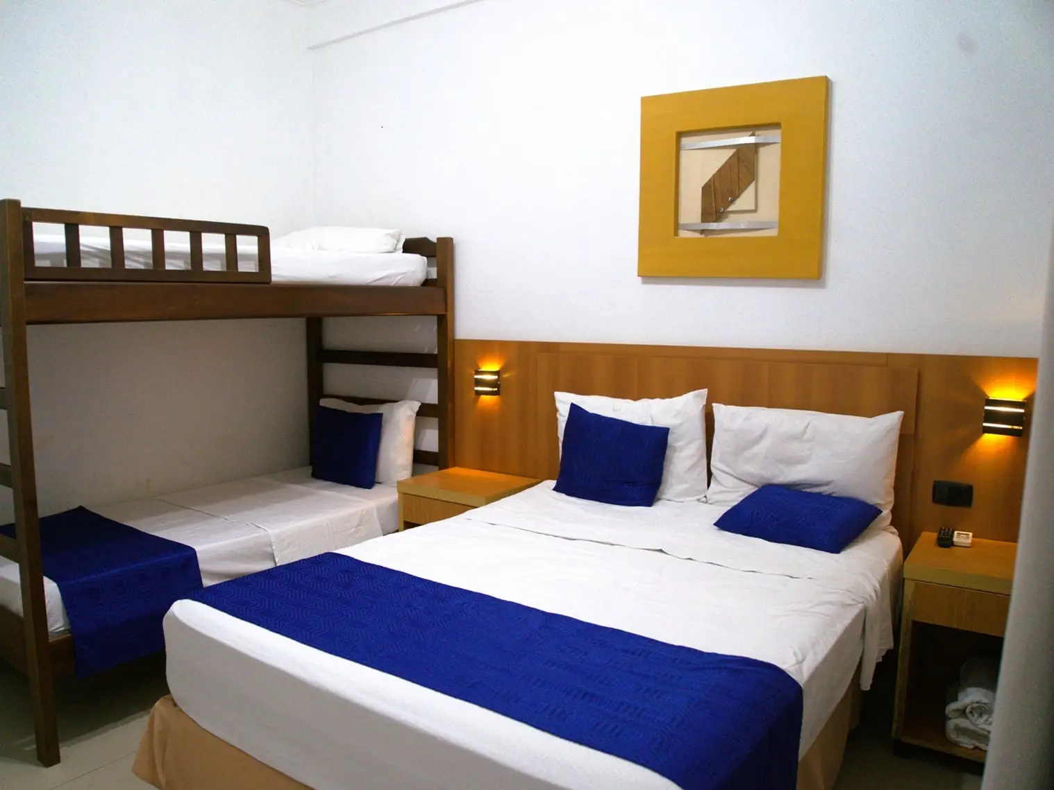 Spacious family hotel room featuring a large double bed and a wooden bunk bed, both dressed in white linens with navy blue accent runners and pillows, set against a white wall with a yellow framed picture.