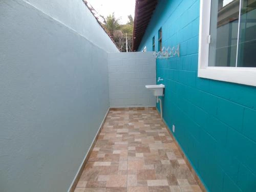 A slim outdoor corridor between two buildings, featuring a light gray textured wall on the left, a bright teal block wall on the right, a small white ceramic wall-mounted sink, a metal coat rack, and a floor covered in light beige and brown square ceramic tiles.
