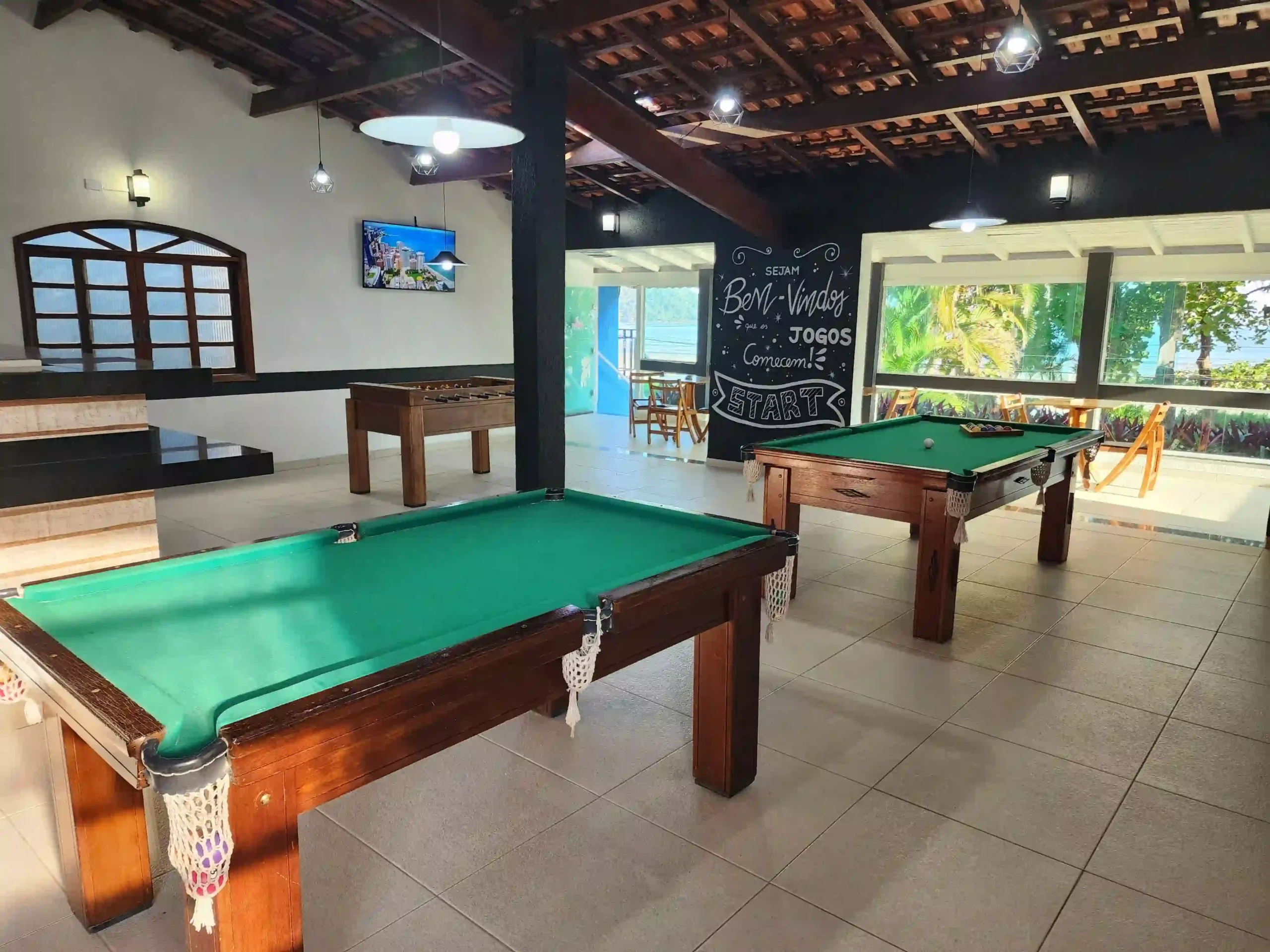 Bright indoor games room with two green felt pool tables and a wooden foosball table, set on tiled flooring beneath a dark exposed wood beam ceiling with pendant lighting and a large window overlooking palm trees.