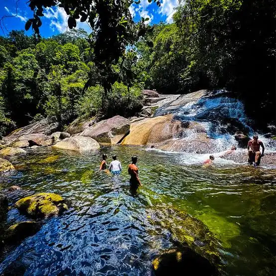 A group of tourists gathered on a sandy, sunny riverbank next to a clear, greenish forest pool. Some people are standing and wading in the shallow water, while others sit on the shore, with backpacks resting nearby.