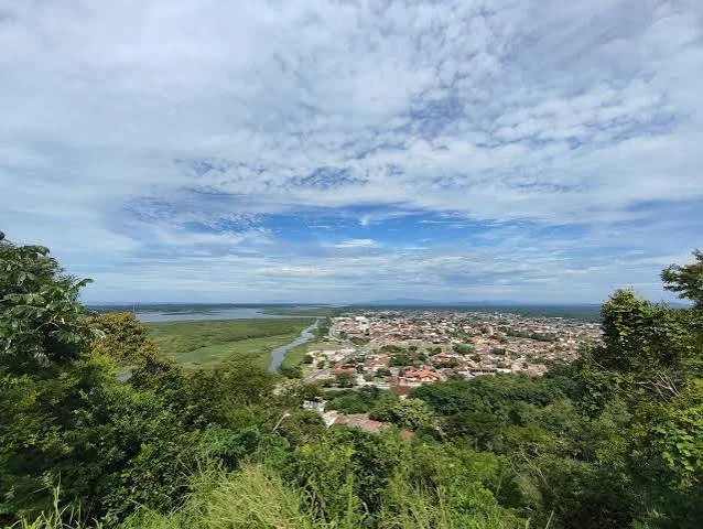 A wide, elevated view looking down from a green, grassy hilltop onto a town laid out in a grid pattern. A wide, dark-colored river or inlet separates the dense town from a distant, flat, green landscape, stretching out toward a hazy horizon under a bright sky.